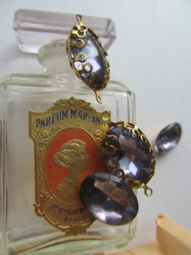 May include: A vintage glass bottle with a gold label that reads "PARFUM MARIANA" and "JN. GIRAUD PARIS". The bottle is decorated with three gold-toned metal embellishments with dark grey glass stones.