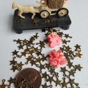 K&ouml;nnte beinhalten: Ein gro&szlig;er Haufen goldener sternf&ouml;rmiger Charms mit Sprungringen. Zwei rosafarbene Plastikblumen mit goldenen Stern-Charms liegen oben auf dem Haufen. Ein Centst&uuml;ck liegt in der Mitte des Haufens.