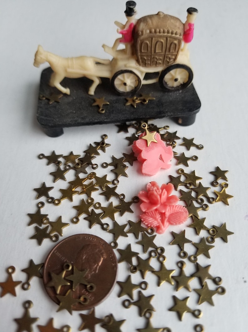 K&ouml;nnte beinhalten: Ein gro&szlig;er Haufen goldener sternf&ouml;rmiger Charms mit Sprungringen. Zwei rosafarbene Plastikblumen mit goldenen Stern-Charms liegen oben auf dem Haufen. Ein Centst&uuml;ck liegt in der Mitte des Haufens.