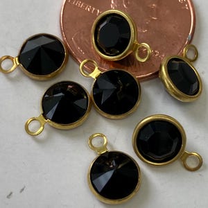 May include: Close-up of several black faceted gemstone charms with gold-colored metal settings and small loops. The charms are round and oval, with a penny in the background for size reference. The gemstones have a shiny, reflective surface.