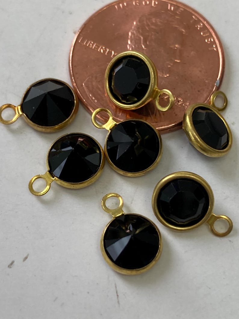 May include: Close-up of several black faceted gemstone charms with gold-colored metal settings and small loops. The charms are round and oval, with a penny in the background for size reference. The gemstones have a shiny, reflective surface.
