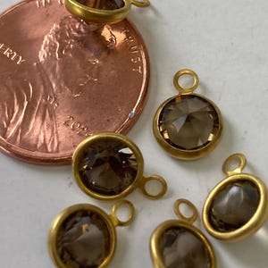 May include: Close-up of several gold-toned bezel-set pendants with smoky quartz stones. Each pendant has a small loop for attaching to a chain or other jewelry component. A U.S. penny is included for size reference.