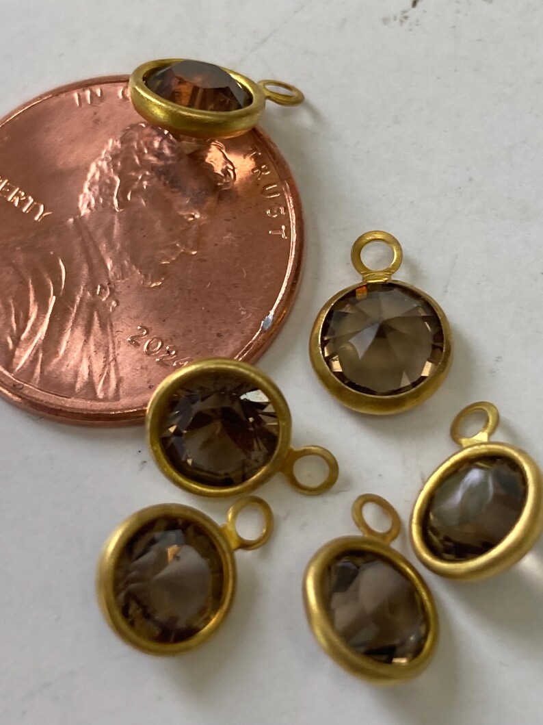May include: Close-up of several gold-toned bezel-set pendants with smoky quartz stones. Each pendant has a small loop for attaching to a chain or other jewelry component. A U.S. penny is included for size reference.