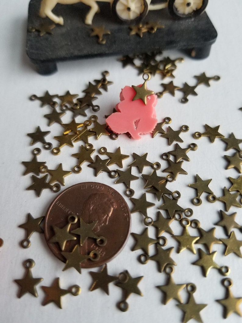 K&ouml;nnte beinhalten: Eine gro&szlig;e Menge kleiner, goldfarbener Metall-Stern-Anh&auml;nger mit einem Sprungring an jedem Stern. Die Sterne sind auf einer wei&szlig;en Oberfl&auml;che verstreut. Ein Cent-St&uuml;ck ist zur Gr&ouml;&szlig;enbestimmung enthalten.