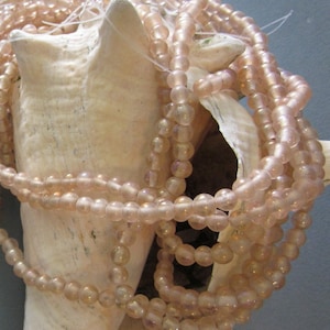 Puede incluir: Primer plano de hebras de cuentas de vidrio rosa claro. Las cuentas son pequeñas y redondas, con un acabado ligeramente iridiscente. Se colocan sobre una gran concha de color blanquecino, creando una exhibición delicada y texturizada.