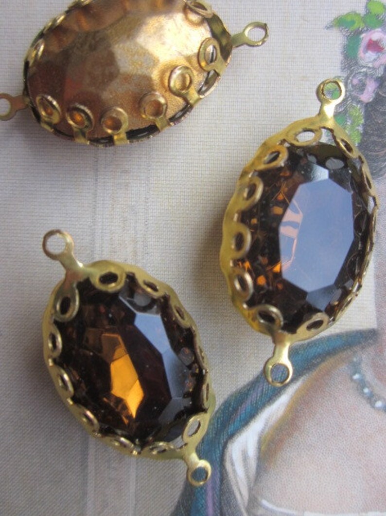 Pode incluir: Tr&ecirc;s engastes de metal dourado com pedras de vidro ovais. As pedras s&atilde;o transparentes com um tom castanho. Os engastes t&ecirc;m uma borda decorativa de pequenos c&iacute;rculos.