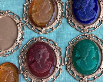 Könnte beinhalten: Acht antike Kameenbroschen mit goldfarbenen Metallrahmen. Die Kameen zeigen unterschiedlich farbiges Glas mit erhabenen Bildern von Frauenprofilen. Die Kameen sind in den Farben Bernstein, Blau, Grün und Rot.