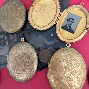 May include: Four oval-shaped gold-toned metal lockets with intricate floral engravings on the exterior. One locket is open, revealing a gold-toned interior frame containing a small black and white portrait photograph of a person wearing a suit.