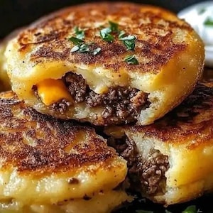 May include: Close-up of golden-brown potato cakes, cut open to reveal a savoury filling. The filling appears to be ground meat and melted cheese. Garnished with fresh green herbs, these savoury cakes are a hearty meal.