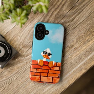 Könnte beinhalten: Blaue iPhone-Hülle mit einer Cartoon-Ente mit Sonnenbrille, die über eine Backsteinmauer blickt. Der Hintergrund ist ein blauer Himmel mit einer weißen Wolke. Die Hülle hat ein glänzendes Finish.