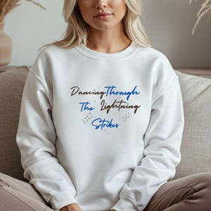 Pode incluir: Camisola branca com o texto "Dancing Through The Lightning Strikers" em azul e castanho. A camisola tem mangas compridas e gola redonda. O texto é rodeado por pequenas estrelas e corações.