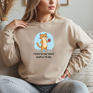 Puede incluir: Sudadera beige con un gato de dibujos animados naranja y blanco sosteniendo una taza de café. El gato está sobre un fondo azul claro. El texto dice: "I have to say weird stuff or I'll die".