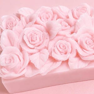 Puede incluir: Una pastilla de jabón rectangular de color rosa claro con una superficie esculpida decorada con múltiples rosas y hojas tridimensionales. Todo el objeto es de un color rosa pastel uniforme con una base sólida y lisa con relieve floral.