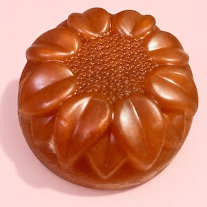 May include: A round, amber-colored soap bar molded into the shape of a sunflower. The center features a textured, dotted pattern representing seeds, surrounded by smooth, raised petals that extend toward the outer edge of the circular form.
