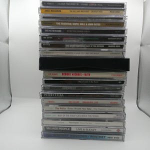 May include: A stack of compact discs with various album titles and artist names visible. The CDs are in clear plastic cases, with a black cassette tape in the middle of the stack. Titles include albums by George Michael, The Police, and Whitney Houston.