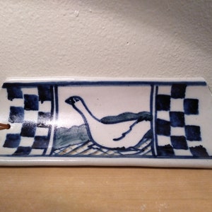 May include: A white ceramic dish with a blue and white checkered border. A blue and white bird is painted in the center of the dish.