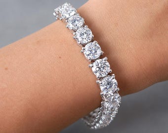 May include: A silver-toned tennis bracelet featuring a continuous row of round, clear, faceted stones set in four-prong metal settings. The bracelet wraps around the wrist, showcasing the reflective quality and uniform size of each stone.