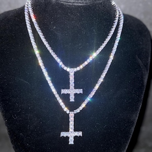 Peut inclure: Deux colliers argentés avec des pendentifs en forme de croix. Les colliers sont composés de petites pierres scintillantes, créant un éclat brillant. Les pendentifs en croix sont également incrustés des mêmes pierres. Les bijoux sont présentés sur une surface en velours noir.