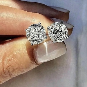 May include: A pair of round-cut stud earrings featuring clear, faceted stones set in a four-prong silver-toned metal setting. The earrings exhibit high clarity and brilliance, reflecting light with multiple facets visible on the surface of each stone.