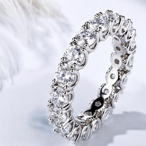 Könnte beinhalten: Ein silberfarbener Eternity-Ring mit einer durchgehenden Reihe runder, im Brillantschliff geschliffener, klarer Steine in einer Krappenfassung. Die Metallschiene ist poliert und die Steine sind so angeordnet, dass sie den Ring vollständig umschließen.
