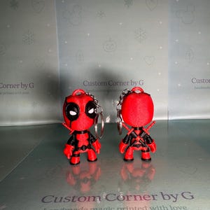 Könnte beinhalten: Zwei rote und schwarze Deadpool-Schlüsselanhänger. Jeder Schlüsselanhänger zeigt eine kleine, 3D-gedruckte Figur der Marvel Comics-Figur, komplett mit Schwertern auf dem Rücken. Die Schlüsselanhänger haben silberne Schlüsselringe. Der Text "Custom Corner by G" ist sichtbar.