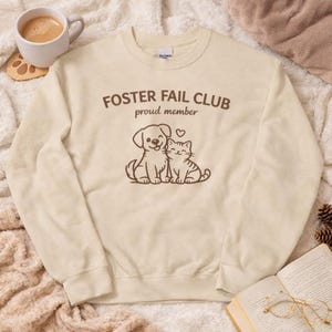 Peut inclure: Sweat-shirt crème avec l'inscription "FOSTER FAIL CLUB proud member" en marron. En dessous, une illustration marron d'un chien et d'un chat. Le sweat-shirt est fait d'une matière douce.