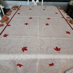 May include: Cream-colored rectangular tablecloth with a fall theme. Features red maple leaves scattered across the surface and a border with pumpkins and sunflowers. A dark red stripe frames the edges.