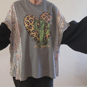 May include: A gray t-shirt with a heart-shaped design featuring a cactus and leopard print. The sleeves are black, and the shoulders have a patterned fabric with beige, pink, and orange accents. The shirt is casual wear.