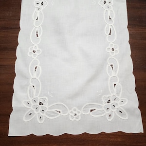 May include: White embroidered table runner with a scalloped edge. The runner features a floral and paisley design along the borders. The embroidery is detailed and delicate, adding a touch of elegance to the textile.