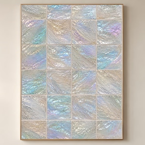 May include: A framed wall art piece featuring a grid of iridescent, textured squares. The squares display a mix of colors including blue, green, and gold, creating a shimmering effect. The artwork is encased in a thin, light-colored frame.