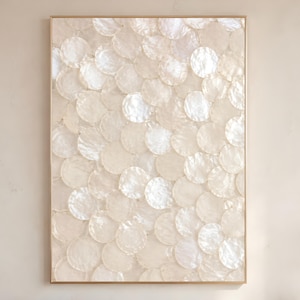 Könnte beinhalten: Ein rechteckiges Wandbild mit einer dichten, überlappenden Anordnung kreisförmiger, irisierender weißer, muschelartiger Scheiben. Das Kunstwerk ist in einen dünnen, goldfarbenen Metallrahmen gefasst und erzeugt eine texturierte, schimmernde Oberfläche.