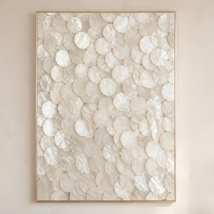 May include: Rectangular wall art featuring a pattern of overlapping, circular, off-white shell pieces. The artwork is framed in a thin, gold-colored frame. The overall aesthetic is neutral and elegant, suitable for various interior styles.