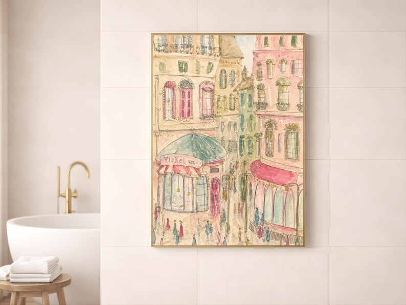 Parisian watercolor art print in a wood frame leaning against a white bathroom wall beside a gold faucet and minimal decor