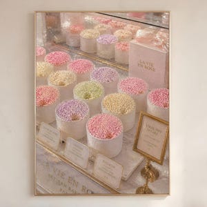 May include: A framed wall art print depicting a bakery display case filled with rows of small, cylindrical cakes topped with colourful sprinkles in shades of pink, yellow, and purple. A box labelled La Vie En Rose and small gold-framed signs are visible.