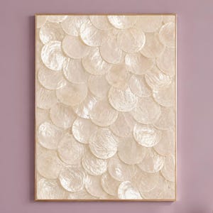 Könnte beinhalten: Ein rechteckiges Wandbild mit einem geschichteten, überlappenden Muster aus kreisförmigen, irisierenden weißen Muschelscheiben. Das Kunstwerk ist in einen dünnen, goldfarbenen Metallrahmen gefasst und erzeugt eine texturierte, reflektierende Oberfläche.