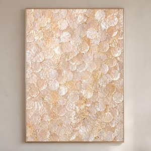 May include: A framed artwork featuring a pattern of overlapping, iridescent, circular shapes in shades of cream, beige, and gold. The artwork has a gold-colored frame and is displayed against a neutral-colored wall. The overall effect is one of texture and subtle shimmer.