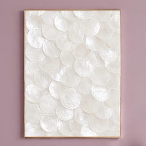 May include: A rectangular wall art piece featuring a vertical arrangement of overlapping, circular white mother-of-pearl discs. The artwork is encased in a thin, metallic gold frame, creating a textured, iridescent surface with a shimmering finish.