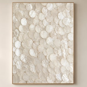 Op de afbeelding: Een ingelijst kunstwerk met een patroon van overlappende, cirkelvormige, iriserende, gebroken witte schelpstukken. Het kunstwerk is ingekaderd in een dunne, lichtbruine houten lijst, waardoor een neutrale en getextureerde wanddecoratie ontstaat.