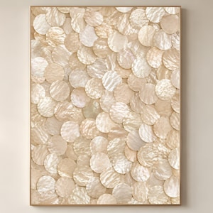 May include: A framed wall art piece featuring a pattern of overlapping, circular, iridescent, mother-of-pearl shells. The shells are in shades of cream and beige, creating a textured, decorative surface. The frame is a light brown color.