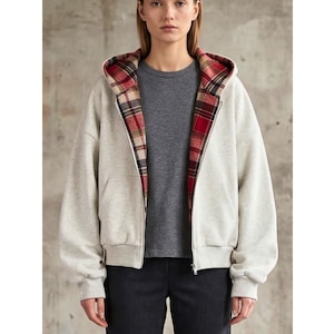 Puede incluir: Una sudadera con capucha gris claro con cremallera y un forro a cuadros rojo, negro y beige en la capucha. La sudadera con capucha tiene una cremallera frontal y dos bolsillos. La persona lleva una camiseta gris y pantalones oscuros.