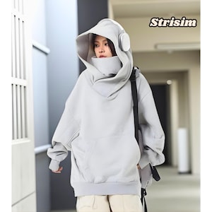 Könnte beinhalten: Ein hellgrauer übergroßer Hoodie mit einem einzigartigen Kapuzendesign und integrierten Kopfhörern. Der Hoodie hat eine große Fronttasche und einen hohen Kragen. Das Wort "Strisim" ist in der oberen rechten Ecke aufgedruckt. Das Model trägt eine schwarze Umhängetasche.