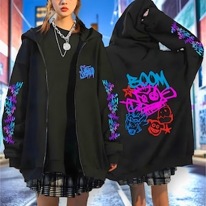 Puede incluir: Sudadera negra con cremallera y gráficos coloridos de estilo grafiti. Las mangas presentan un diseño repetitivo, mientras que la parte trasera muestra la palabra "BOOM" y personajes de dibujos animados en tonos vibrantes. La sudadera tiene capucha y cremallera.