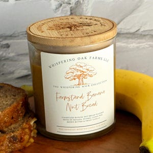 May include: A glass jar candle with a wooden lid, labeled "Farmstand Banana Nut Bread." The label features a tree logo and text. A slice of banana bread and a banana are in the background. The candle is a soy blend.