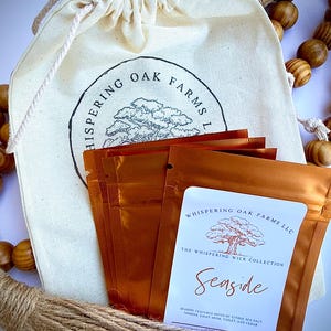 May include: A cream-colored drawstring fabric pouch with the black text Whispering Oak Farms LLC and a tree illustration, containing several metallic copper-colored packets labeled Seaside with notes of citrus, sea salt, jasmine, light musk, violet, and cedar.