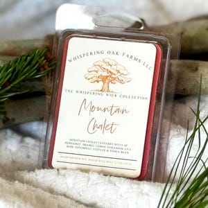 May include: A red wax melt in a clear plastic clamshell. The label reads "Mountain Chalet" and lists fragrance notes. The product is from "Whispering Oak Farms LLC" and is part of "The Whispering Wick Collection".