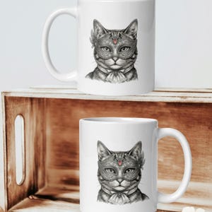 May include: White ceramic mug with a detailed illustration of a cat wearing a masquerade mask. The cat is in grayscale, with the mask decorated with feathers and a red jewel. The mug has a curved handle.