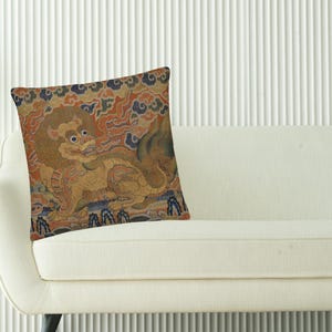 May include: Square decorative pillow with a detailed illustration of a mythical creature in shades of gold, brown, and blue. The pillow is placed on a white sofa, adding a touch of cultural art to the home decor.