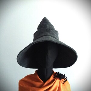 May include: A dark gray witch hat with a wide brim and a pointed top. A black spider is pinned to the orange fabric draped over a black mannequin head. The hat is a Halloween costume accessory.
