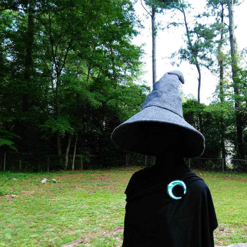 May include: A person wearing a large, gray witch hat and a black cloak. A green and blue crescent moon brooch is pinned to the cloak. The background features a green lawn and trees.