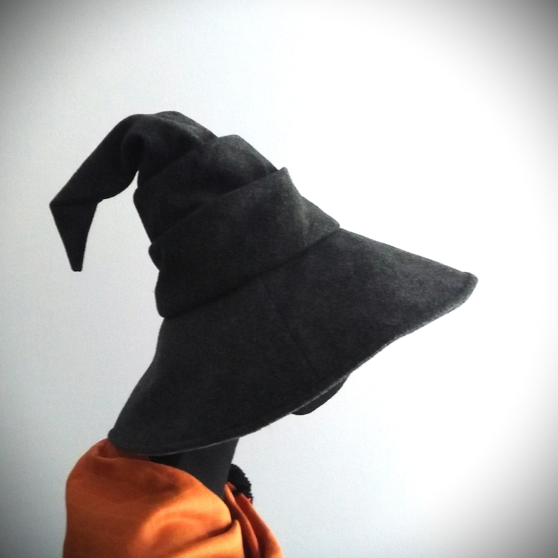May include: A dark gray witch hat with a wide brim and a pointed tip. The hat is made of a soft, felt-like material and has a folded band around the base. An orange scarf is visible beneath the hat.